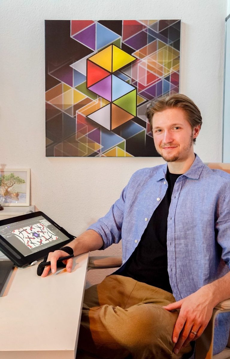 Paul Alexander Heigel Der Künstler sitzt an einem Schreibtisch mit Tablet und vor einem bunten geometrischen Wandbild.